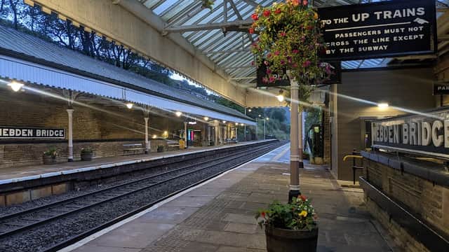 Hebden Bridge bus stop with a covered platform, flower baskets, and a sign directing to up trains. Quiet, early evening scene.