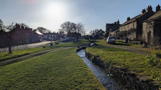 Cherry Tree Cottage alongside a grassy area and stream, with stone houses and trees under a bright sky in Bellerby.