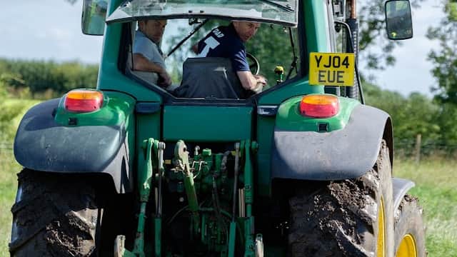 Two men in a green tractor on a dirt path, surrounded by lush greenery and blue skies in Yorkshire.