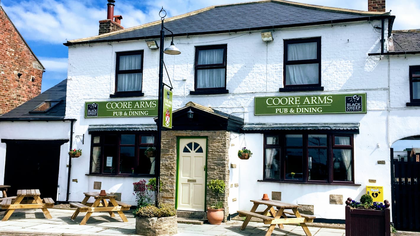 Coore Arms venue with a white facade, green signage, outdoor seating, and a welcoming entrance in Scruton, Yorkshire.
