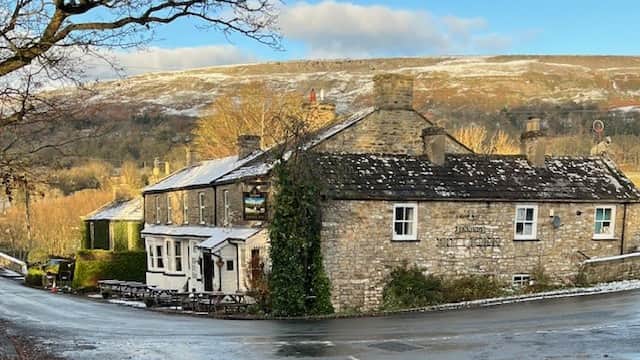 Charming stone hotel with a sloped roof, surrounded by trees and hills, under a blue sky with scattered clouds.