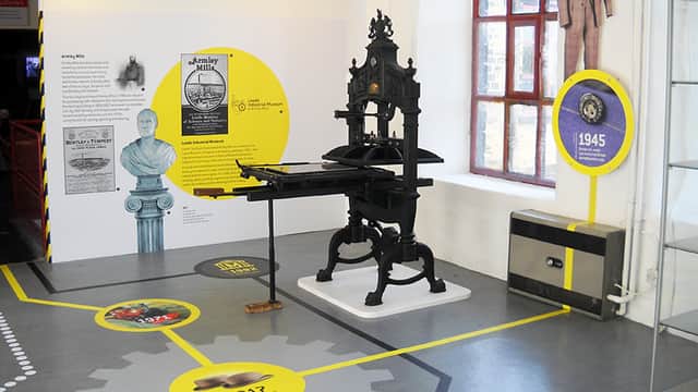 Vintage printing press displayed in a gallery with informational panels and a timeline on the floor.