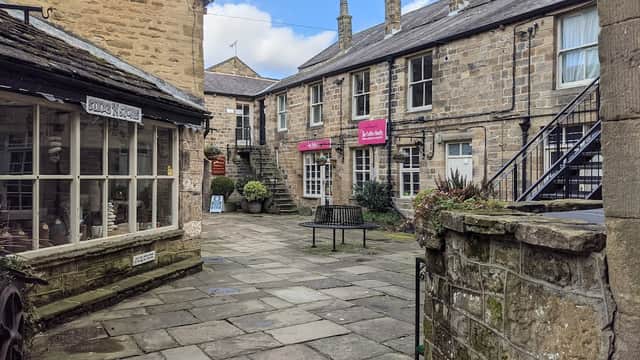 Quaint stone buildings with a courtyard, benches, and a staircase, featuring shops and a blue sky above.