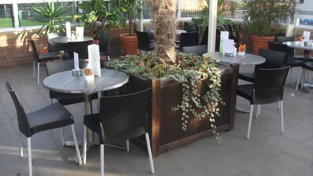Indoor seating area with potted plants, tables, and chairs in a bright, glass-enclosed space.