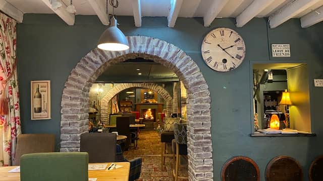 Cozy bar interior with exposed brick arches, wooden beams, a clock, and warm lighting, featuring a fireplace and seating.