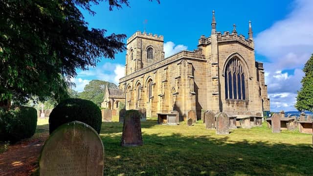 Historic stone church with tall tower, surrounded by gravestones and lush greenery under a bright blue sky.