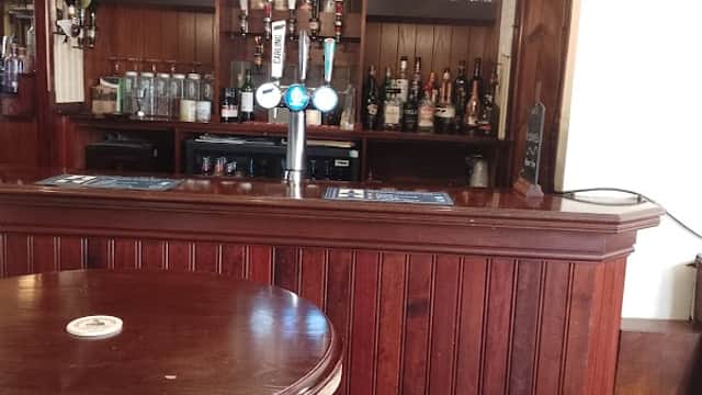 Wooden bar with beer taps, shelves of bottles behind, and a round table with a single stool in a cozy pub setting.