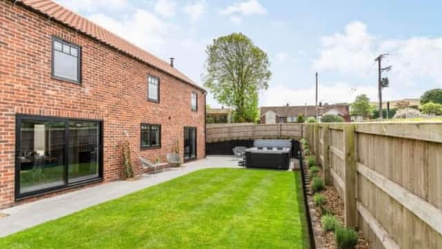 Brick cottage with large windows, manicured lawn, and a hot tub surrounded by wooden fencing and trees.