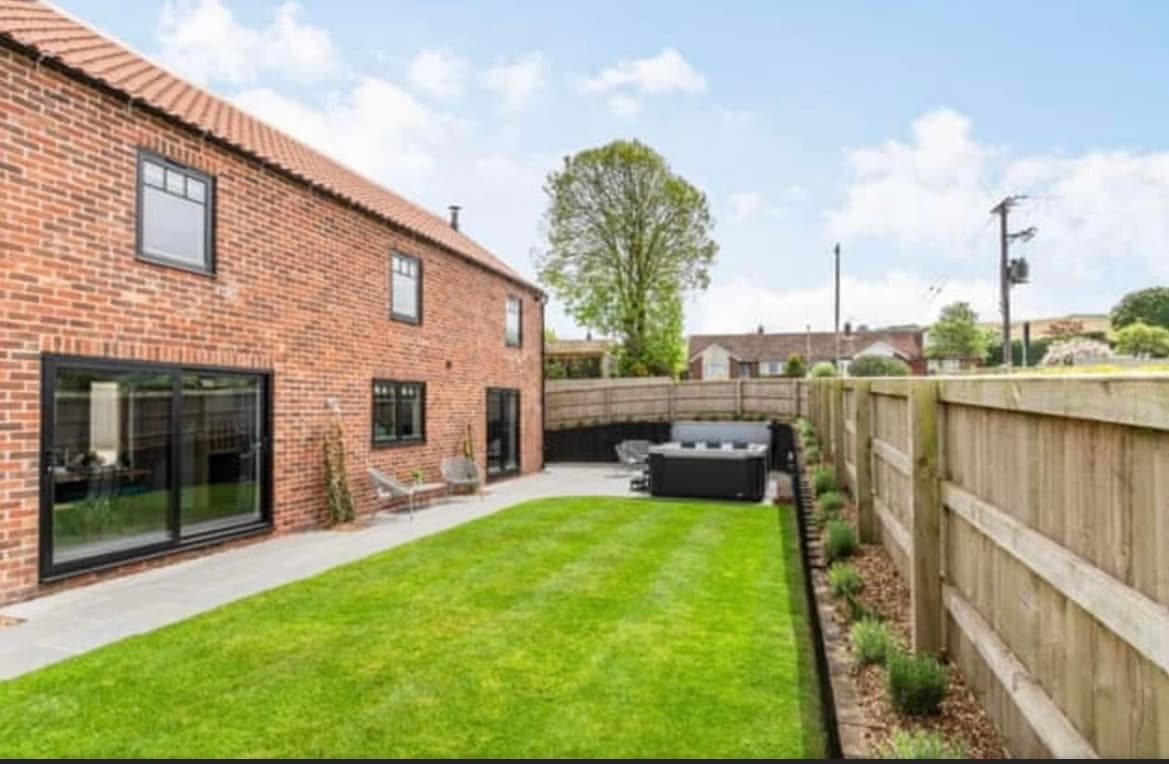Brick cottage with large windows, manicured lawn, and a hot tub surrounded by wooden fencing and trees.