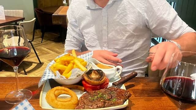 A man in a white shirt and glasses sits at a table with steak, fries, onion rings, and red wine in a cozy restaurant.