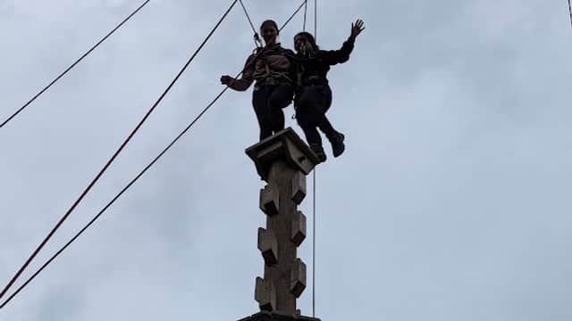Two people standing atop a tall wooden climbing structure against a cloudy sky, with ropes and pulleys visible.