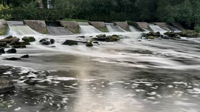 Serene water flowing over a weir, surrounded by lush greenery and smooth stones along the riverbank.