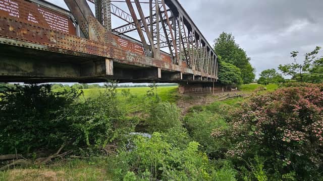 Rusty iron bridge spanning a lush green landscape, surrounded by trees and wildflowers under a cloudy sky.