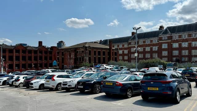 Busy car park filled with parked vehicles, surrounded by brick buildings and blue sky with scattered clouds.