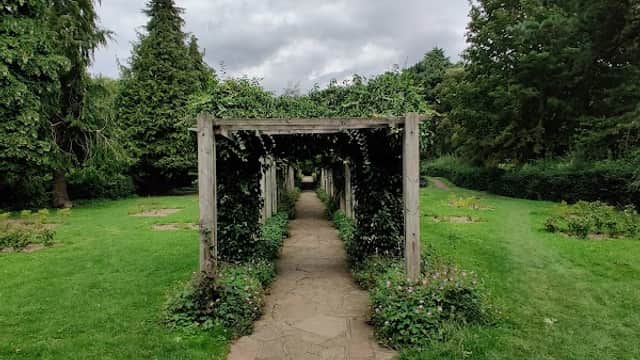 A stone pathway leads through a green park with wooden trellises covered in greenery under a cloudy sky.