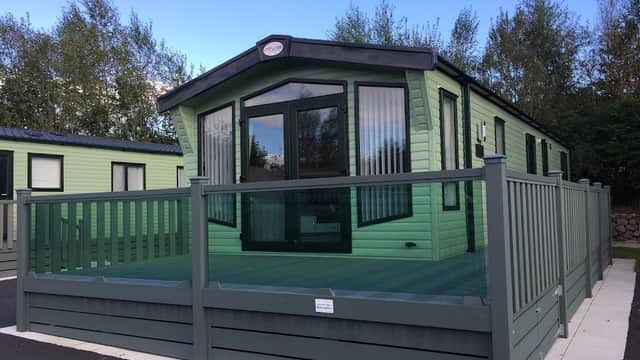 Green caravan with a deck, surrounded by trees, in Langcliffe Caravan Park, Yorkshire. Clear blue sky above.