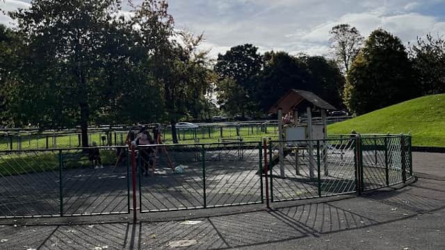Playground with a fenced area, a play structure, and children playing under a sunny sky with scattered clouds.