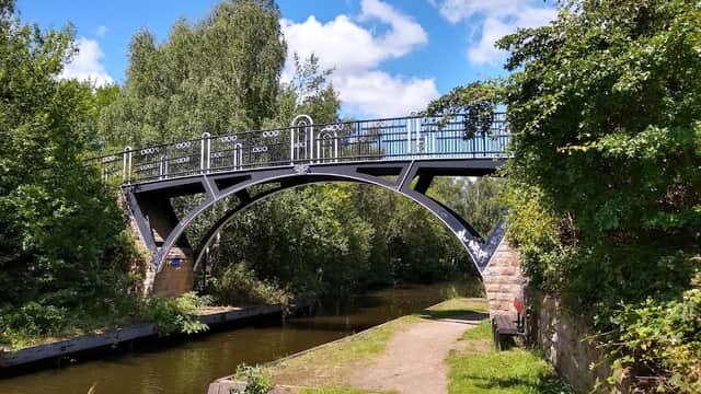 Black iron bridge arching over a calm canal, surrounded by lush greenery and a walking path.