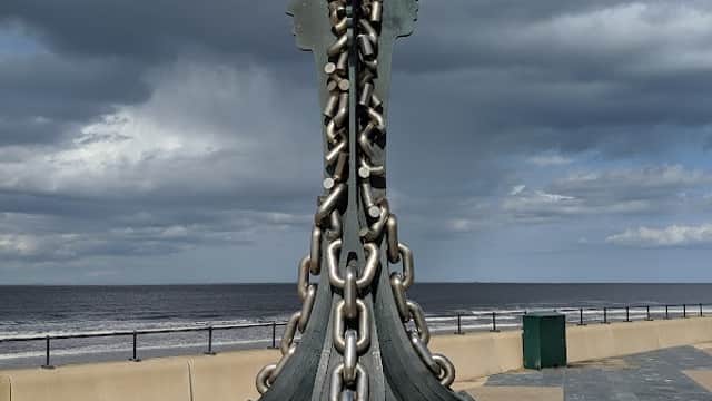 Sculpture resembling chains and blades on a coastal promenade with a cloudy sky and sea in the background.
