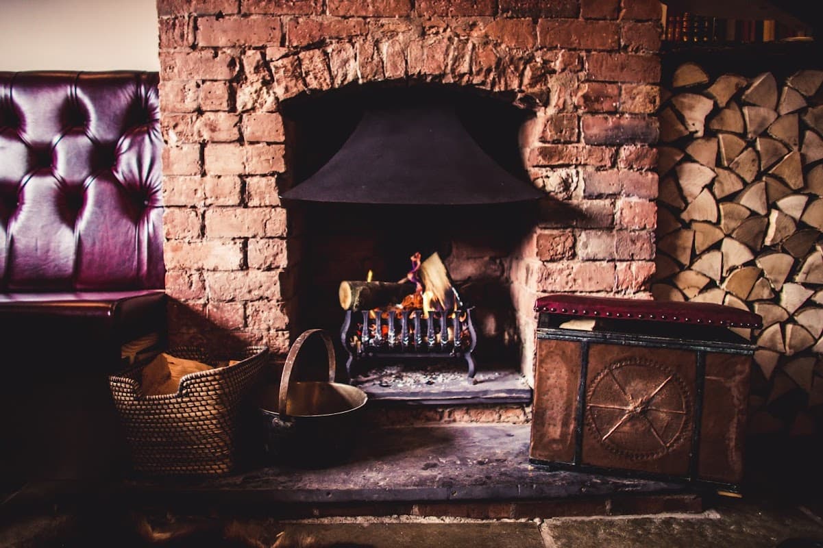 Cozy fireplace with a black hood, wooden logs stacked beside it, and a vintage chest in a warm, inviting interior.