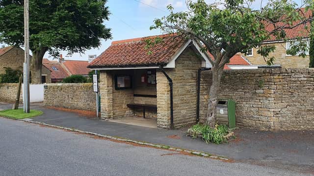 Hutton Buscel bus shelter with a stone wall, bench, and nearby greenery along a quiet street in Yorkshire.