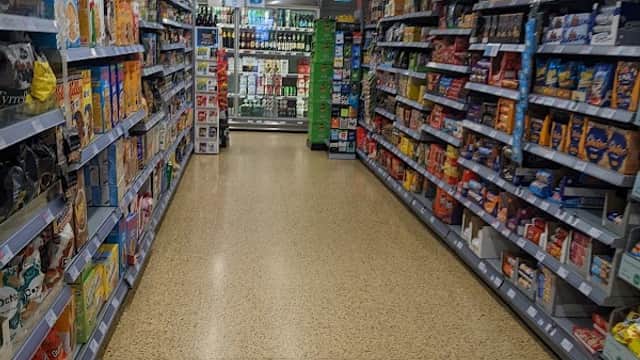 Aisle in a grocery store lined with shelves of various food products, leading to a checkout area in the background.