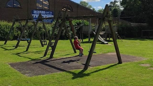 A playground with swings on grass, a child sitting on a swing, and a building in the background under a cloudy sky.