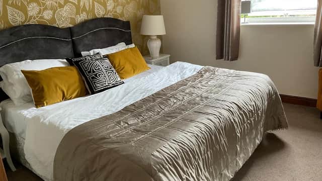 Cozy bedroom with a large bed, decorative pillows, bedside lamp, and patterned wallpaper in a cottage setting.