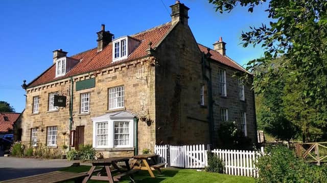 Historic stone pub with a red-tiled roof, white windows, and a green sign, surrounded by lush greenery and picnic tables.