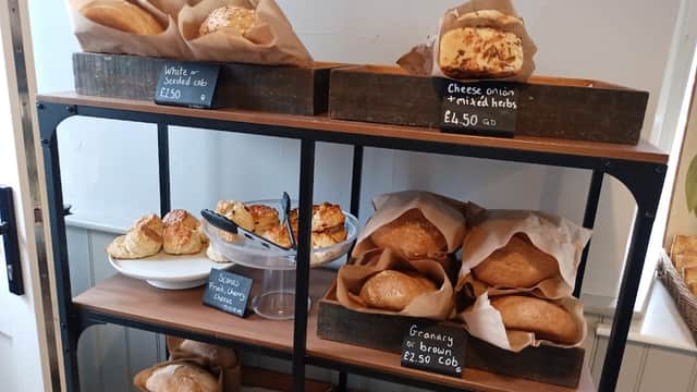 Wooden shelves display various baked goods, including loaves, rolls, and scones, with prices on chalkboard signs.