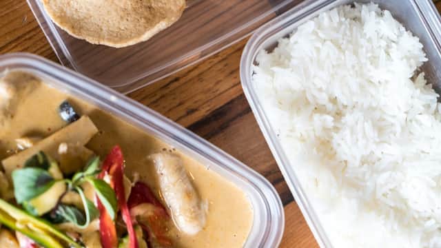 Thai curry with vegetables, served with white rice and crispy snacks in takeaway containers on a wooden surface.