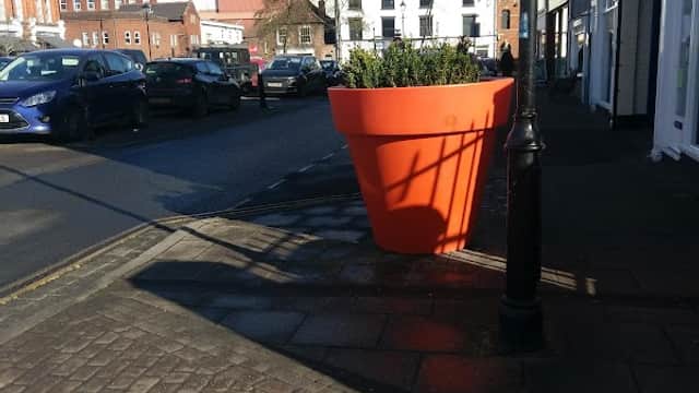 Bright orange flower pot on a cobblestone sidewalk, with cars parked and buildings in the background under a clear blue sky.