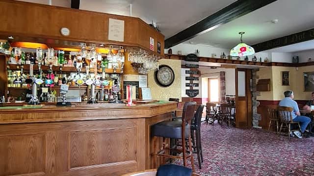 Cozy pub interior featuring a wooden bar, colorful drinks, floral decor, and patrons seated at tables on patterned carpet.