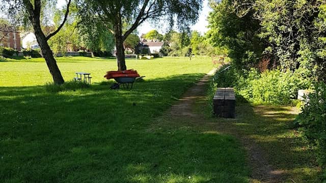 Lush green park with trees, a pathway, benches, and a picnic table under clear blue skies in South Milford.