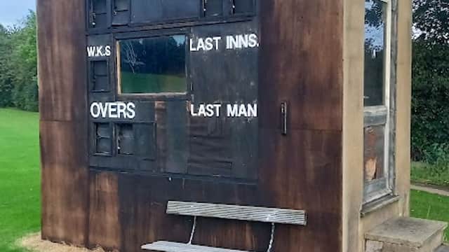 Weathered scoreboard building at Raskelf Cricket Club, with benches and a clock, set on a green cricket field.