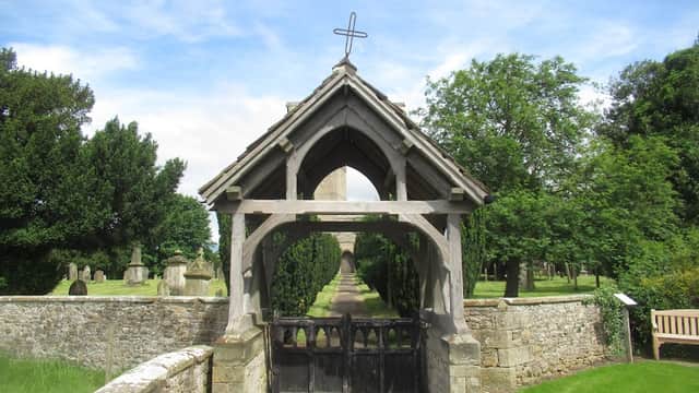 Wooden entrance with a cross, leading to a church surrounded by trees and gravestones in a serene landscape.