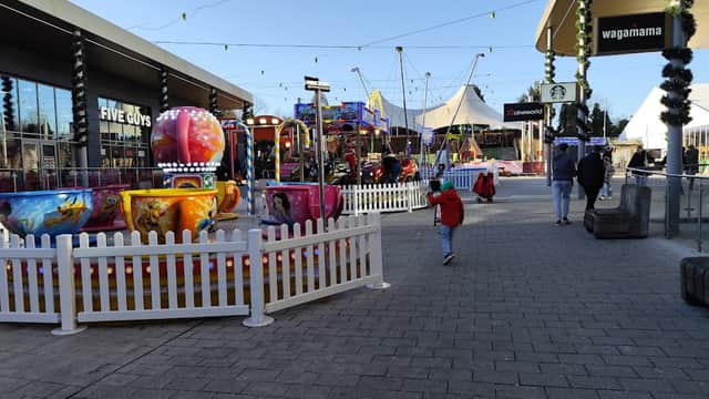 Colorful amusement rides and attractions in a festive car park area, with shoppers walking and string lights above.