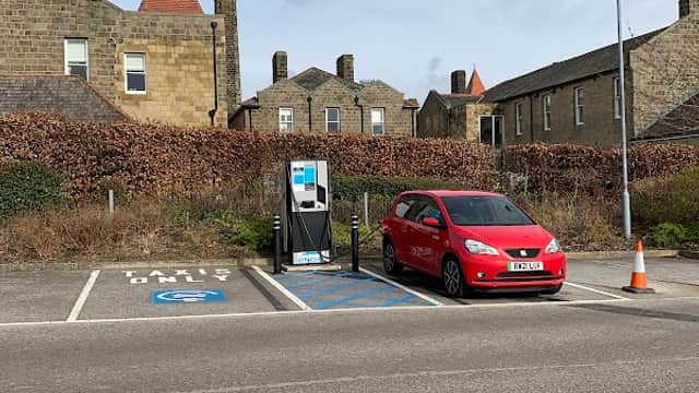 Parking area with a red car and an electric vehicle charging station, surrounded by stone buildings and greenery.