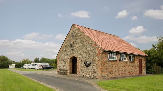 Stone building with a red-tiled roof, surrounded by green grass and parked caravans under a cloudy sky.
