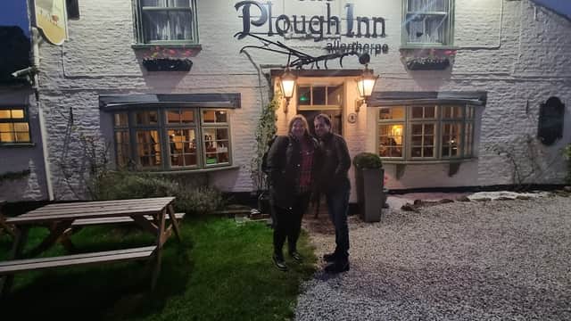 Two women stand outside The Plough Inn, a charming pub with a gravel path and lanterns, surrounded by greenery.