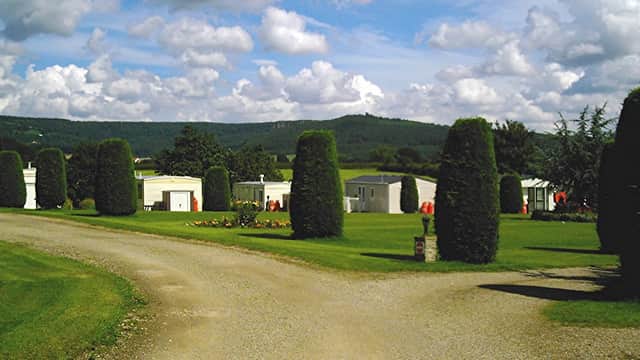 Gravel path leads through manicured lawns and tall hedges, with holiday cabins nestled against a backdrop of rolling hills.