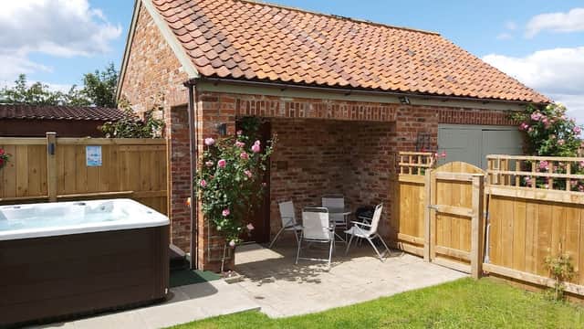 Charming brick cottage with a red-tiled roof, hot tub, and patio seating, surrounded by greenery and wooden fencing.