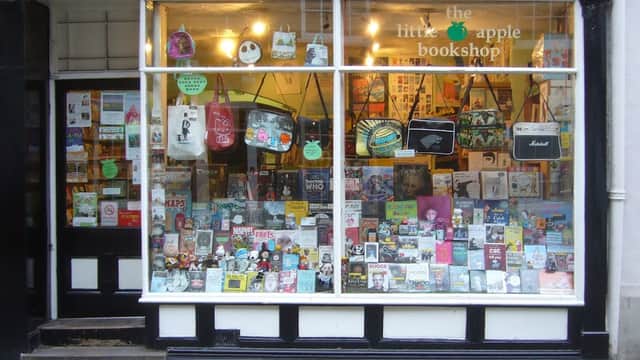 Colorful window display of "The Little Apple Bookshop" featuring various books, bags, and decorative items.
