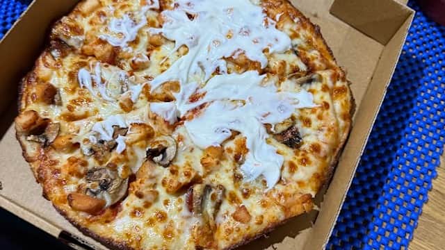 A pizza with cheese, chicken, and a drizzle of sauce in an open pizza box on a blue textured surface.