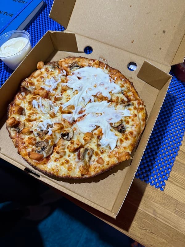 A pizza with cheese, chicken, and a drizzle of sauce in an open pizza box on a blue textured surface.