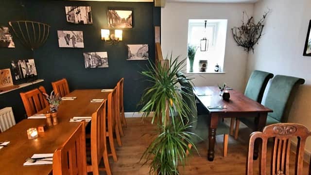 Cozy bar interior with wooden tables, chairs, plants, and black wall featuring historical photos.