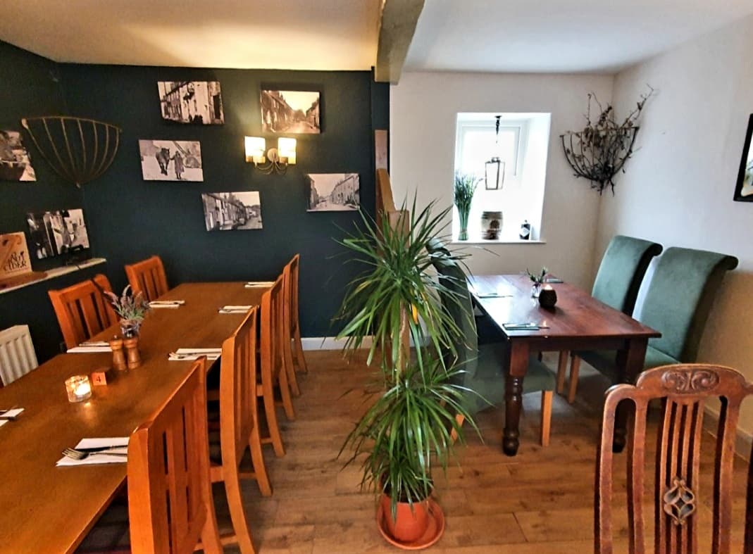 Cozy bar interior with wooden tables, chairs, plants, and black wall featuring historical photos.
