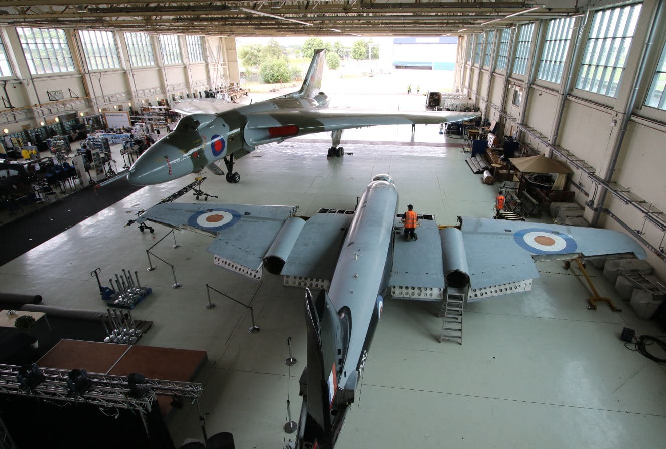 Two vintage military aircraft in a large hangar, with technicians working and equipment scattered around.