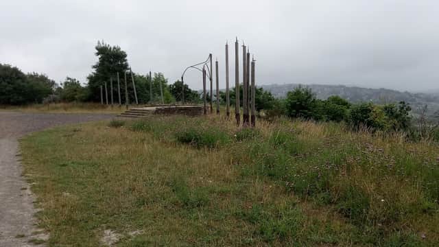 Grass-covered path leading to tall metal sculptures amidst greenery and a misty landscape.