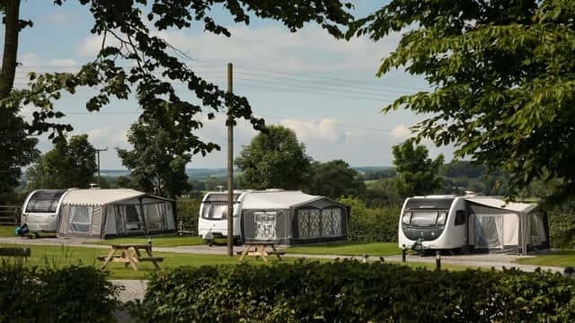Caravans set in a lush green landscape, surrounded by trees and distant hills under a clear blue sky.